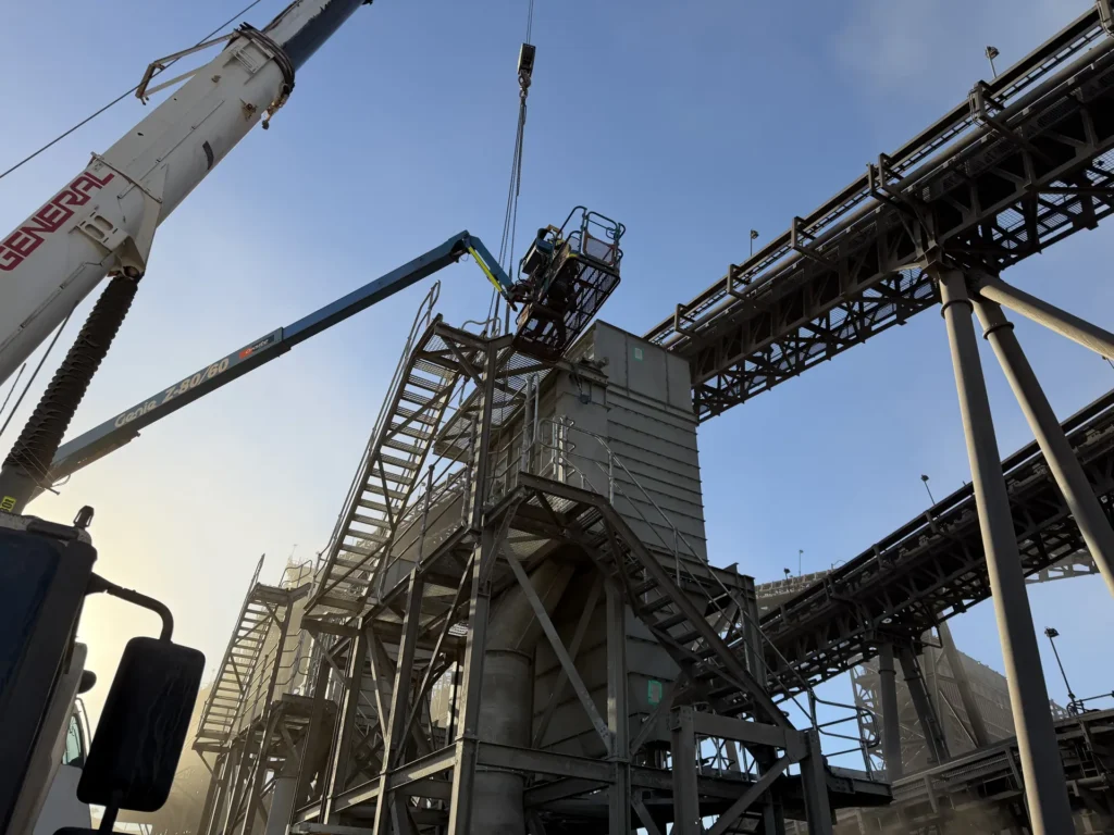 Workers in an EWP basket at a conveyor transfer tower with a crane overhead